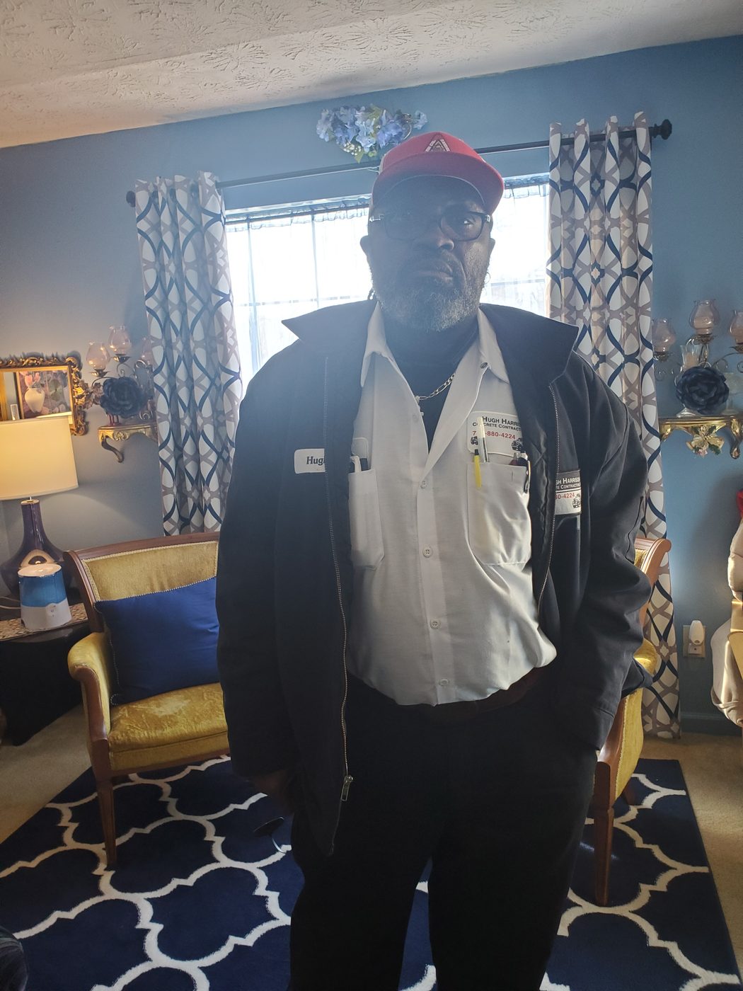 Hugh Harrison, Jr. in his Hugh Harrison Concrete Contracting work shirt, red cap, and glasses
