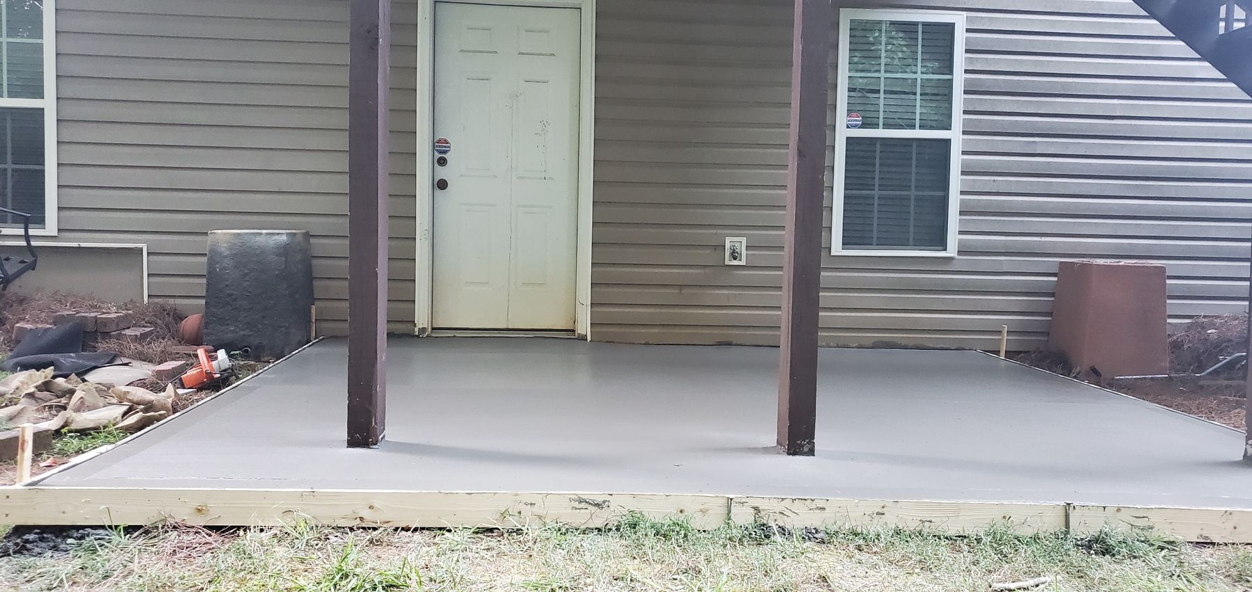 Freshly finished concrete patio slab still in the wooden forms, set around two porch posts